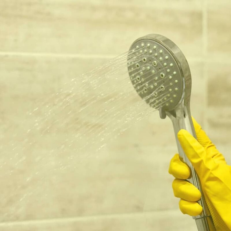 A person wearing yellow rubber gloves sprays a handheld shower head against a tiled bathroom wall to rinse away soap scum and mold-causing moisture.