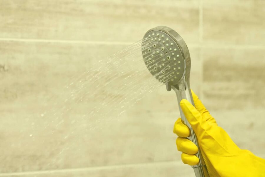 A person wearing yellow rubber gloves sprays a handheld shower head against a tiled bathroom wall to rinse away soap scum and mold-causing moisture.