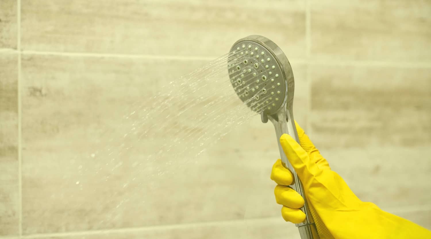 A person wearing yellow rubber gloves sprays a handheld shower head against a tiled bathroom wall to rinse away soap scum and mold-causing moisture.
