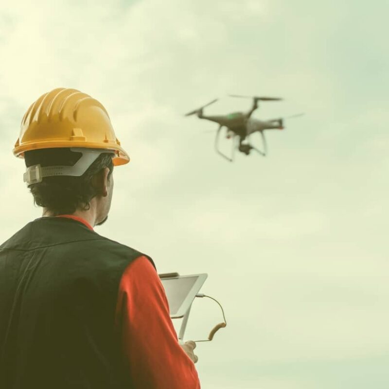 A professional wearing a yellow hard hat and safety vest looks up at a drone flying in a cloudy sky while operating a tablet controller.