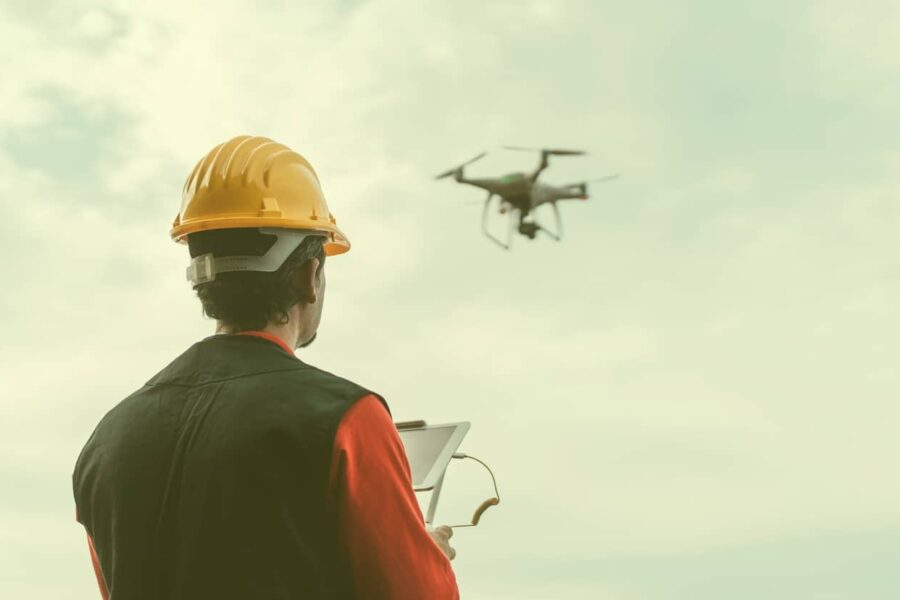 A professional wearing a yellow hard hat and safety vest looks up at a drone flying in a cloudy sky while operating a tablet controller.